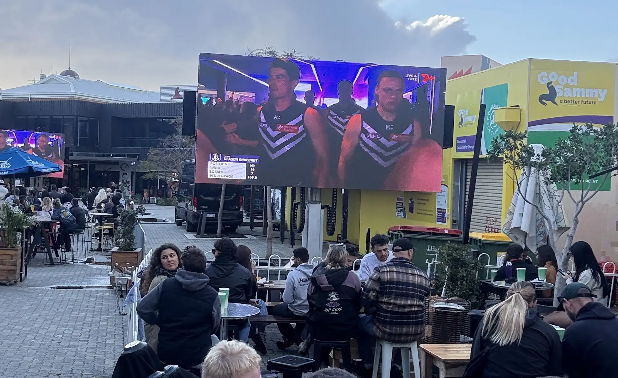 Patrons at an outdoor dining area watching a sports broadcast on a large LED screen