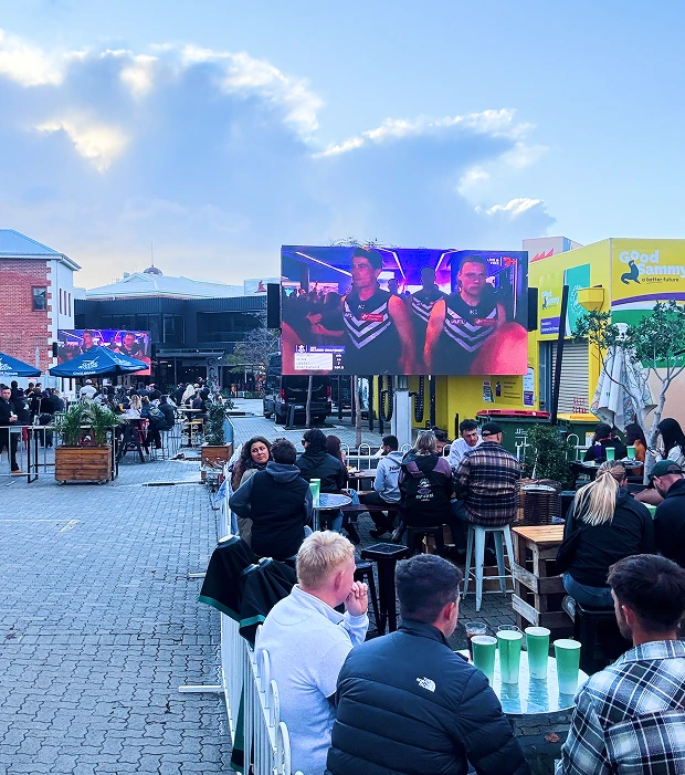 Outdoor dining courtyard with patrons watching a sports broadcast on a large LED screen