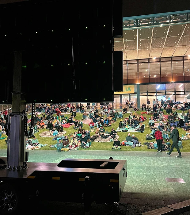 Crowd seated on a lawn at an evening outdoor event in front of a stage